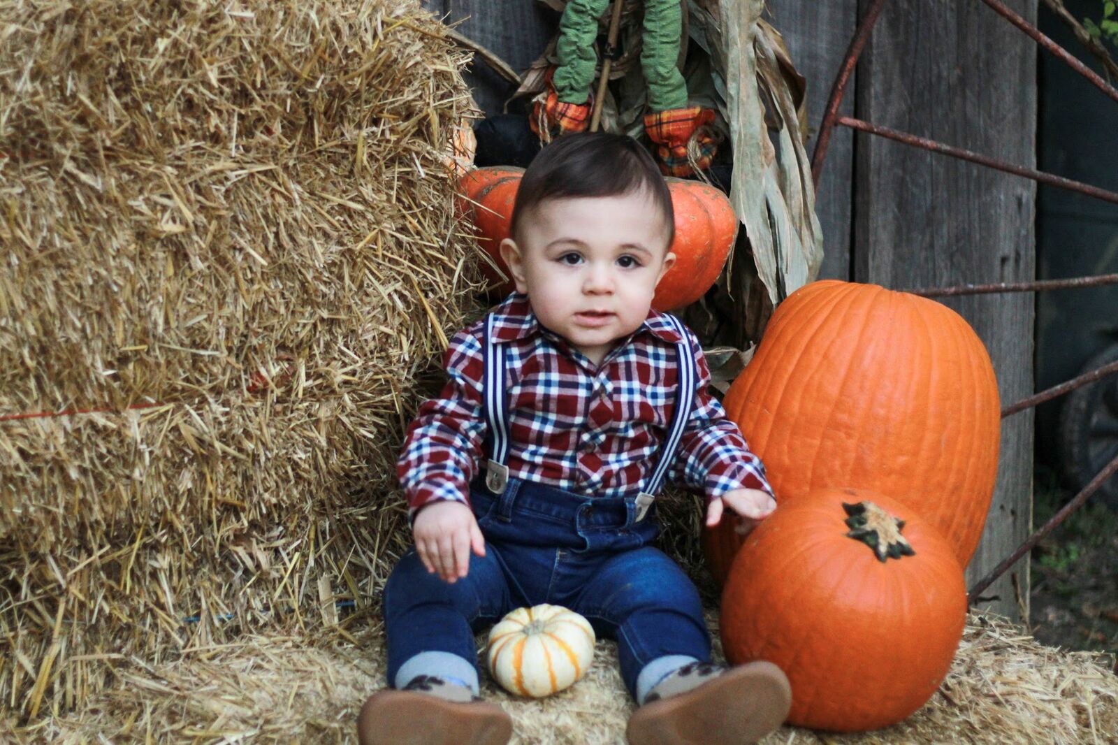 A baby in a plaid shirt sitting on a hay bale beside pumpkins